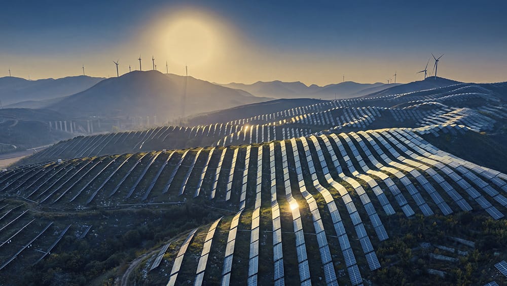 Vogelperspektive einer hügeligen Landschaft mit Solarmodulen und Windkrafträdern am Horizont.